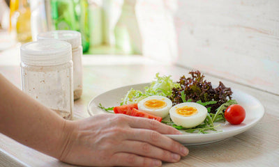 A fair-skinned hand resting near a plate of eggs tomato and greens with a plain supplement jar