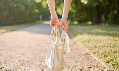 Hands carrying a canvas tote and a glass water bottle along a sunlit park path
