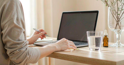 Fair-skinned hands working at a sunlit desk with a laptop, water glass and a liver support dropper
