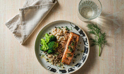 Overhead flat lay of a plate with salmon, white beans, broccoli and farro on a pale oak table