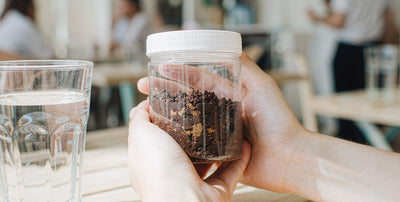 Fair-skinned hands holding a plain unlabeled desiccated liver jar and water on a sunny cafe table.
