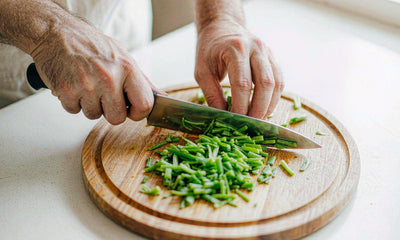 Hands chopping fresh green vegetables on a round wooden board in a sunlit kitchen, seen close up