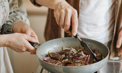 Fair-skinned hands of two people searing liver with onions at a stove, no faces visible, warm light