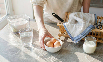 Fair-skinned hand in a knit sleeve placing a glass of water and a bowl of eggs on a sunlit kitchen counter