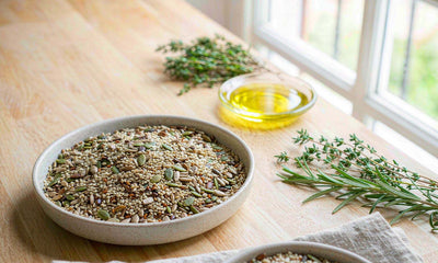 Bowl of mixed seeds with a small dish of golden olive oil and fresh herbs on a light wooden counter