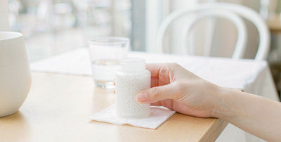 A fair-skinned hand holding a plain supplement bottle at a calm cafe table by a bright window