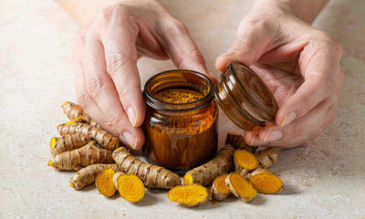 Hands holding an amber jar of ground turmeric beside fresh turmeric root slices on pale stone