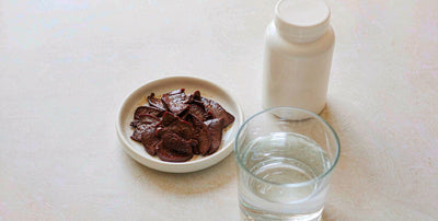 A flat-lay of cooked beef liver slices beside a glass of water and a plain unlabeled supplement jar