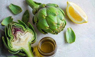 Overhead flat lay of a halved globe artichoke with lemon and a small amber jar on pale marble