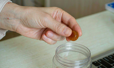 Macro of a fair-skinned hand placing a supplement gummy from a plain jar on a bright desk beside a laptop.