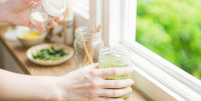 Fair-skinned hands holding a glass jar of greens on a bright windowsill, no head or face in the frame