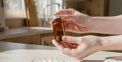 Fair-skinned hands holding a plain unlabeled supplement bottle on a bright kitchen counter in morning light.