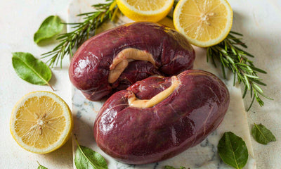 Raw beef kidney on a light marble board with fresh rosemary, lemon halves, and green leaves