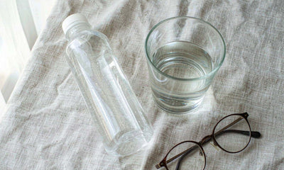 A plain unlabeled bottle beside a glass of water and reading glasses on a light linen cloth
