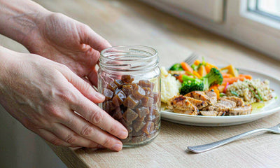 Macro of fair-skinned hands placing a plain beef-liver gummy jar beside a wholesome meal plate, no face shown.