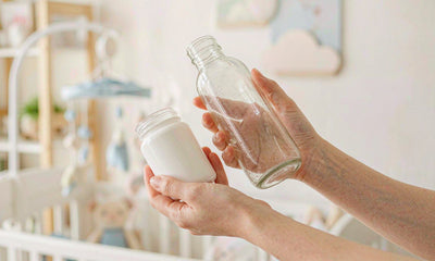 Fair-skinned hands holding a water bottle and a plain supplement jar in a bright soft nursery-toned room