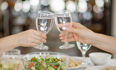 Two pairs of fair-skinned hands clinking water glasses at a bright home dinner table with a salad bowl.