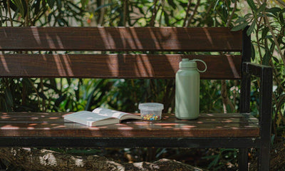 A reusable water bottle and a small plain supplement jar on a sunny park bench beside a book for a calm reset.