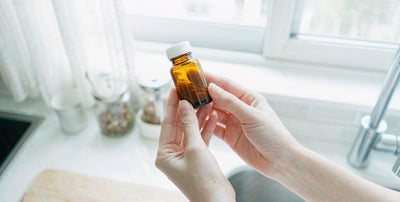 Fair-skinned hands holding a plain supplement bottle in a bright airy kitchen with soft morning light