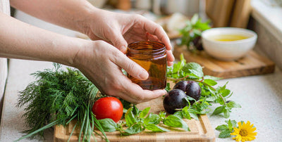 Fair-skinned hands holding a plain amber jar on a wooden board with herbs, nutrient-dense whole food
