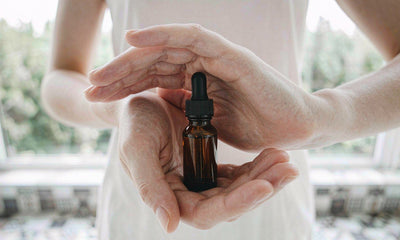 Fair-skinned hands cupped around a small blank amber dropper bottle against a blurred bright kitchen window.