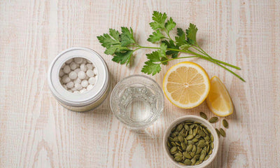 Ingredient flat lay of a capsule tin, water glass, parsley, halved lemon and pumpkin seeds on oak