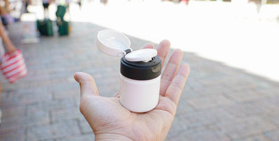 A hand holding a plain white supplement bottle on a sunny street, bright airy on-the-go scene