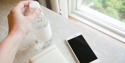 A fair-skinned hand holding a water bottle beside a passport and a phone on a hotel desk by a bright window