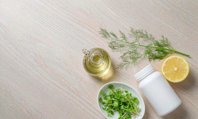 Overhead flat lay of a plain supplement bottle with lemon, olive oil, dill and watercress on pale oak