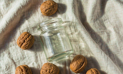 Top-down flat-lay of whole walnuts and a plain glass jar on light linen with soft natural shadows.
