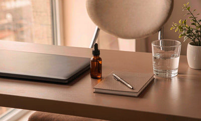 Tidy desk with a small amber dropper bottle, laptop, notebook and water glass in morning light