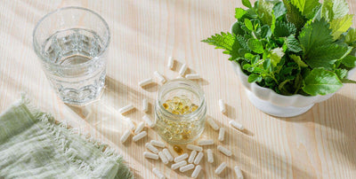 Overhead flat-lay of a glass of water, supplement capsules, and leafy greens on light wood