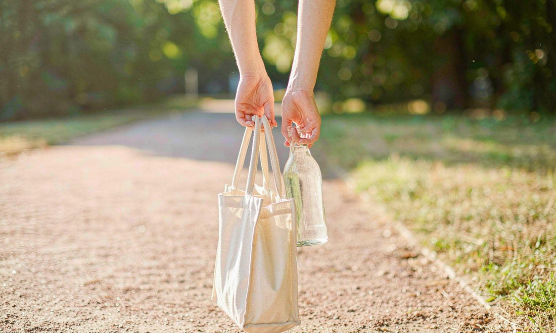Hands carrying a canvas tote and a glass water bottle along a sunlit park path