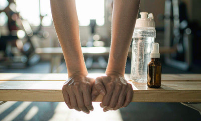 Fair-skinned hands resting on a gym bench beside a plain dropper bottle and water, steady training support
