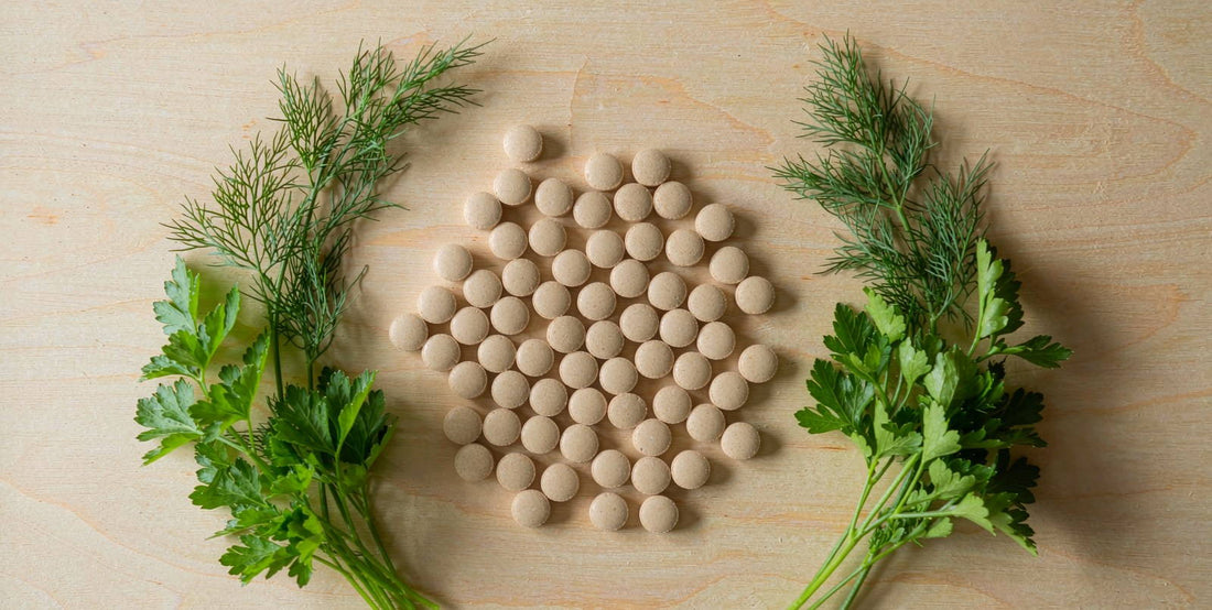 Flat lay of plain supplement pills with fresh green herbs on a bright light wood table in daylight