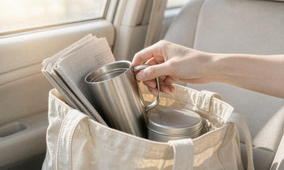 Fair-skinned hand reaching into a canvas tote bag with a travel mug in morning sunlight