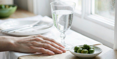A fair-skinned hand resting near a glass of water and olives on a light table after a dinner gathering