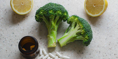 Overhead flat-lay of lemon halves, broccoli, and a plain unlabeled jar with capsules on stone