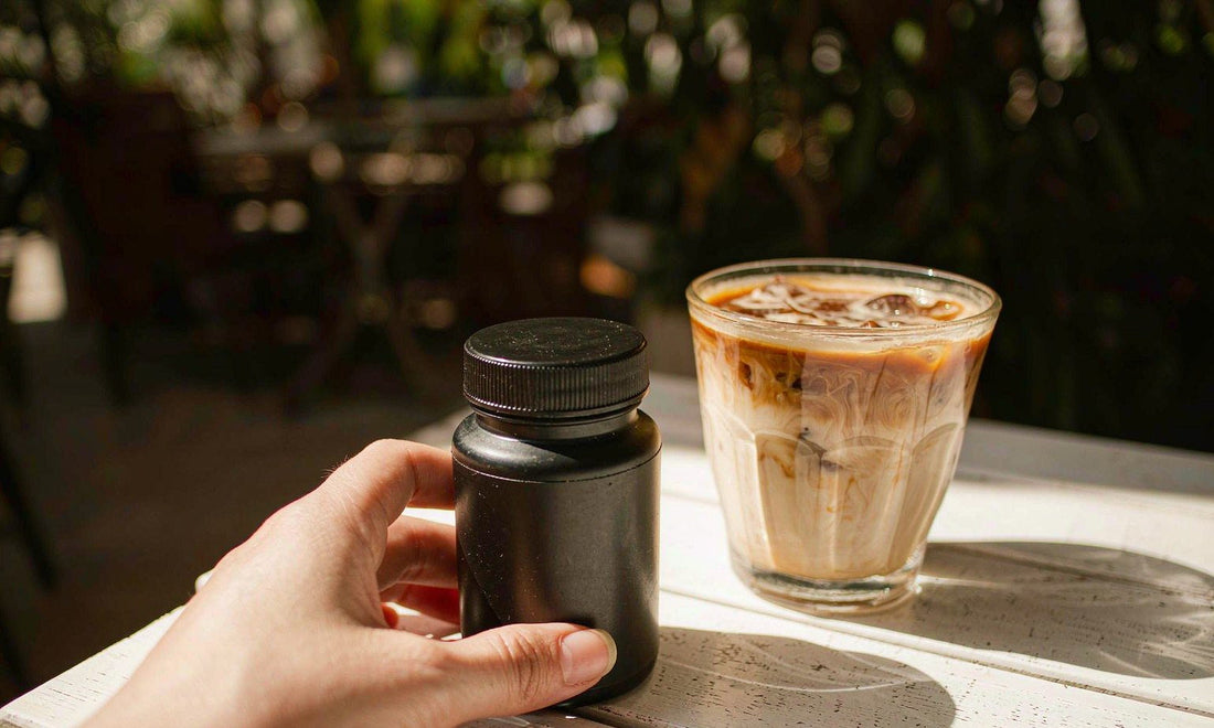 A fair-skinned hand holding a plain unlabeled bottle beside an iced coffee on a sunny cafe table
