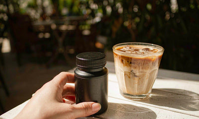 A fair-skinned hand holding a plain unlabeled bottle beside an iced coffee on a sunny cafe table