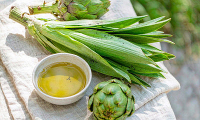 Fresh green artichokes and leaves with a small ceramic dish of olive oil on pale linen cloth