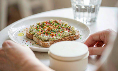 Fair-skinned hands beside a supplement tin, water glass and avocado toast on a sunlit cafe table