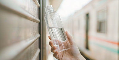 A fair-skinned hand holding a water bottle beside a train window on a shift-change morning