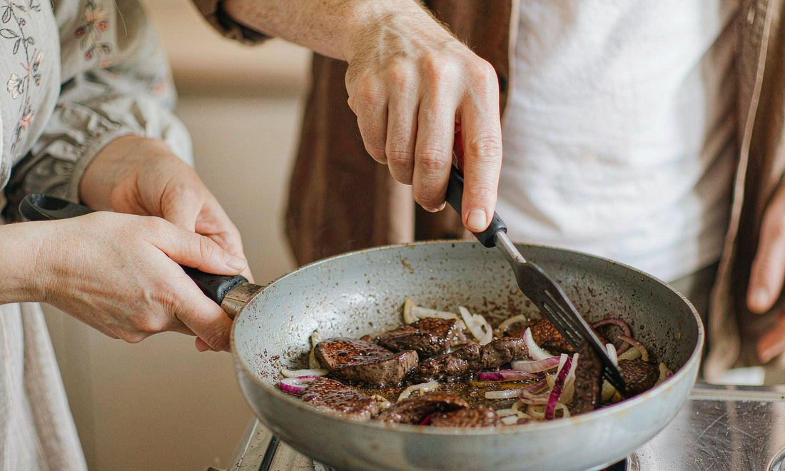 Fair-skinned hands of two people searing liver with onions at a stove, no faces visible, warm light