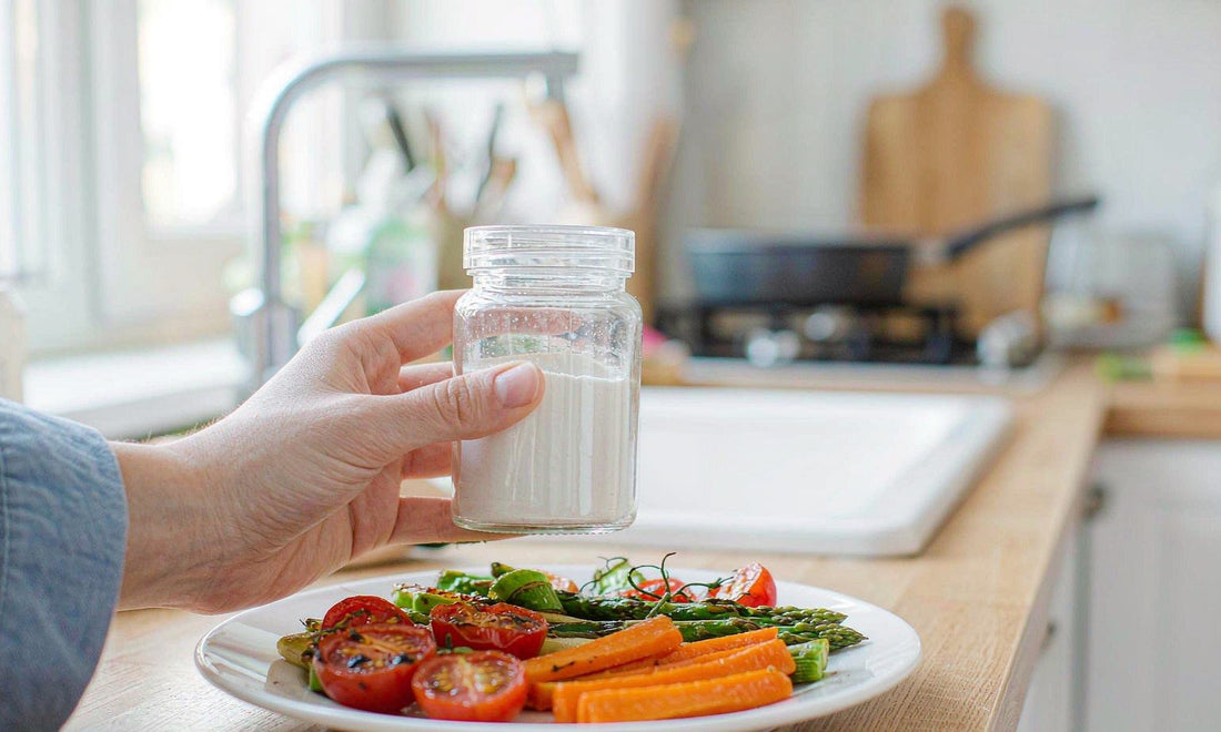 A fair-skinned hand holding a plain glass jar beside a plate of roasted vegetables in a bright kitchen