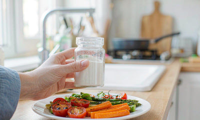 A fair-skinned hand holding a plain glass jar beside a plate of roasted vegetables in a bright kitchen