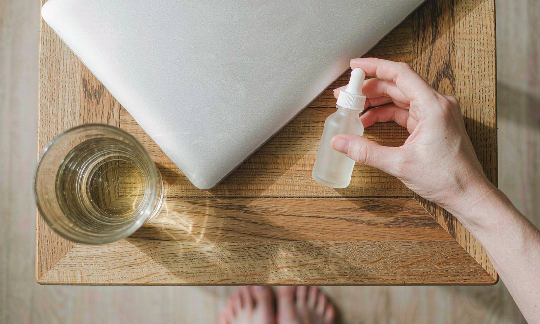 A fair-skinned hand holding a plain dropper bottle on a wooden desk beside a glass of water
