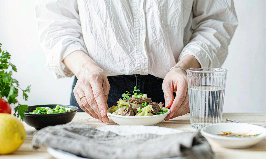 Hands serving a bowl of cooked beef liver with a glass of water on a bright kitchen table