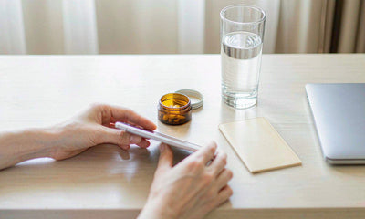 A hotel desk flat lay with an amber supplement tin, water glass and key card in morning light, hands only.