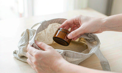Hands placing a plain amber tin into a canvas tote bag on a light desk in soft daylight